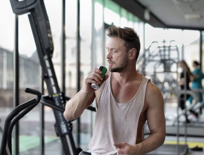 Muscular young sportsman in casual sportswear drinking fresh energy beverage near window while resting after intense workout in modern fitness club