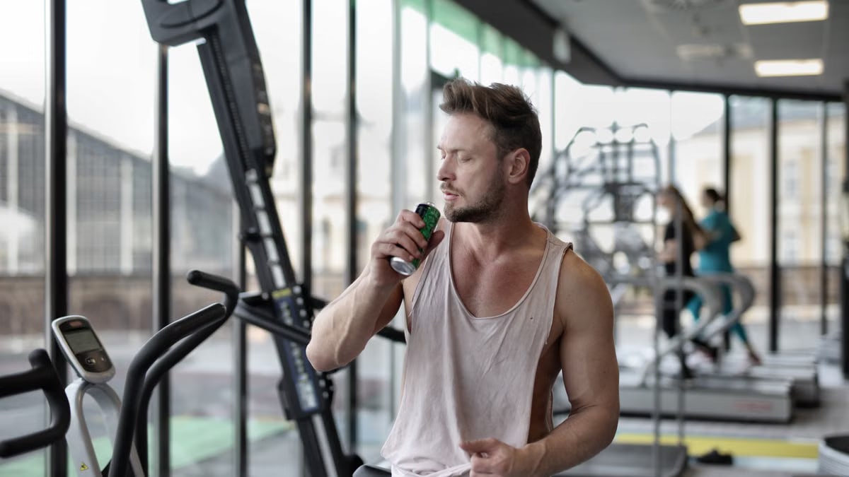 Muscular young sportsman in casual sportswear drinking fresh energy beverage near window while resting after intense workout in modern fitness club